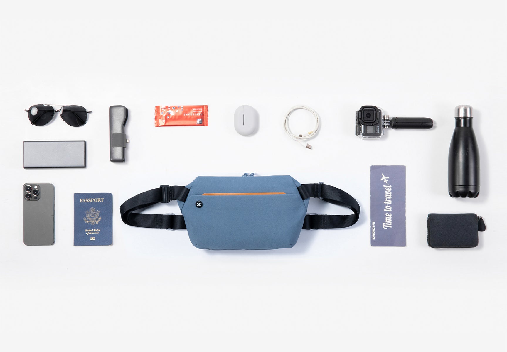 Wanderer Sling in blue color with water bottle, passport, phone, airpod, sunglasses and more that are placed on the table