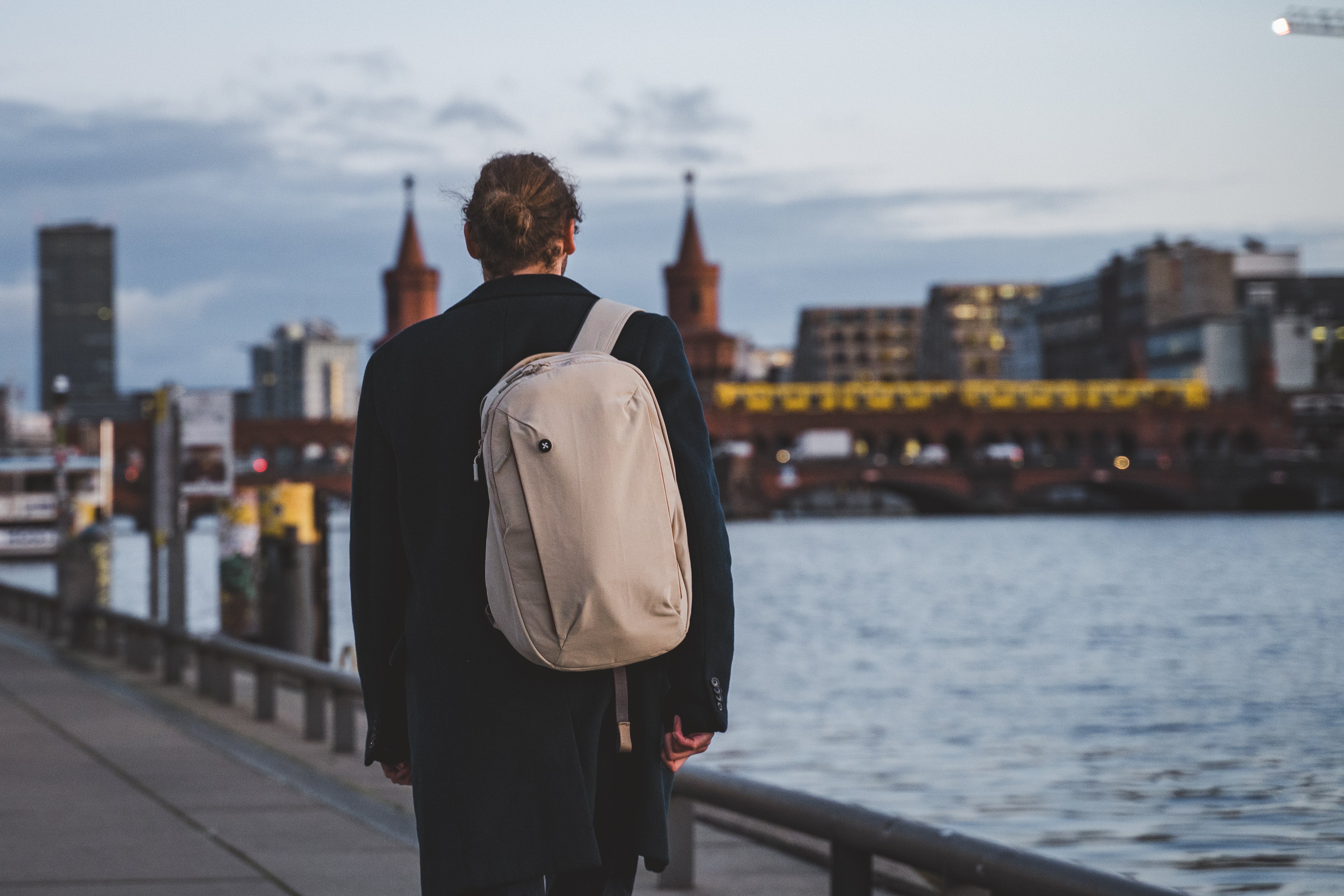 A person wearing a beige mopak backpack, walking along a riverside promenade at dusk with Berlin’s iconic Oberbaum Bridge in the background.