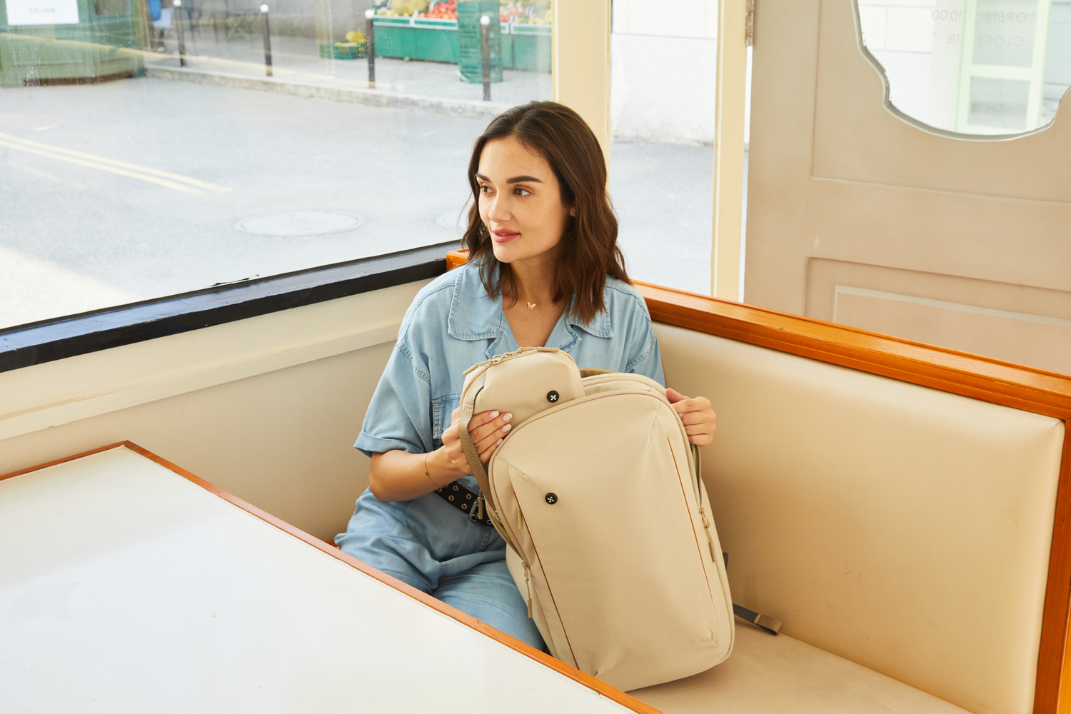 A young woman sitting inside a retro café booth, smiling while holding a Mopak backpack and matching pouch, showcasing an elegant and casual travel look.