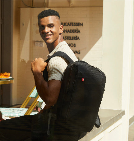 A smiling young man seated at an outdoor café, wearing the black City Adventure Backpack on one shoulder, capturing a relaxed urban lifestyle vibe.
