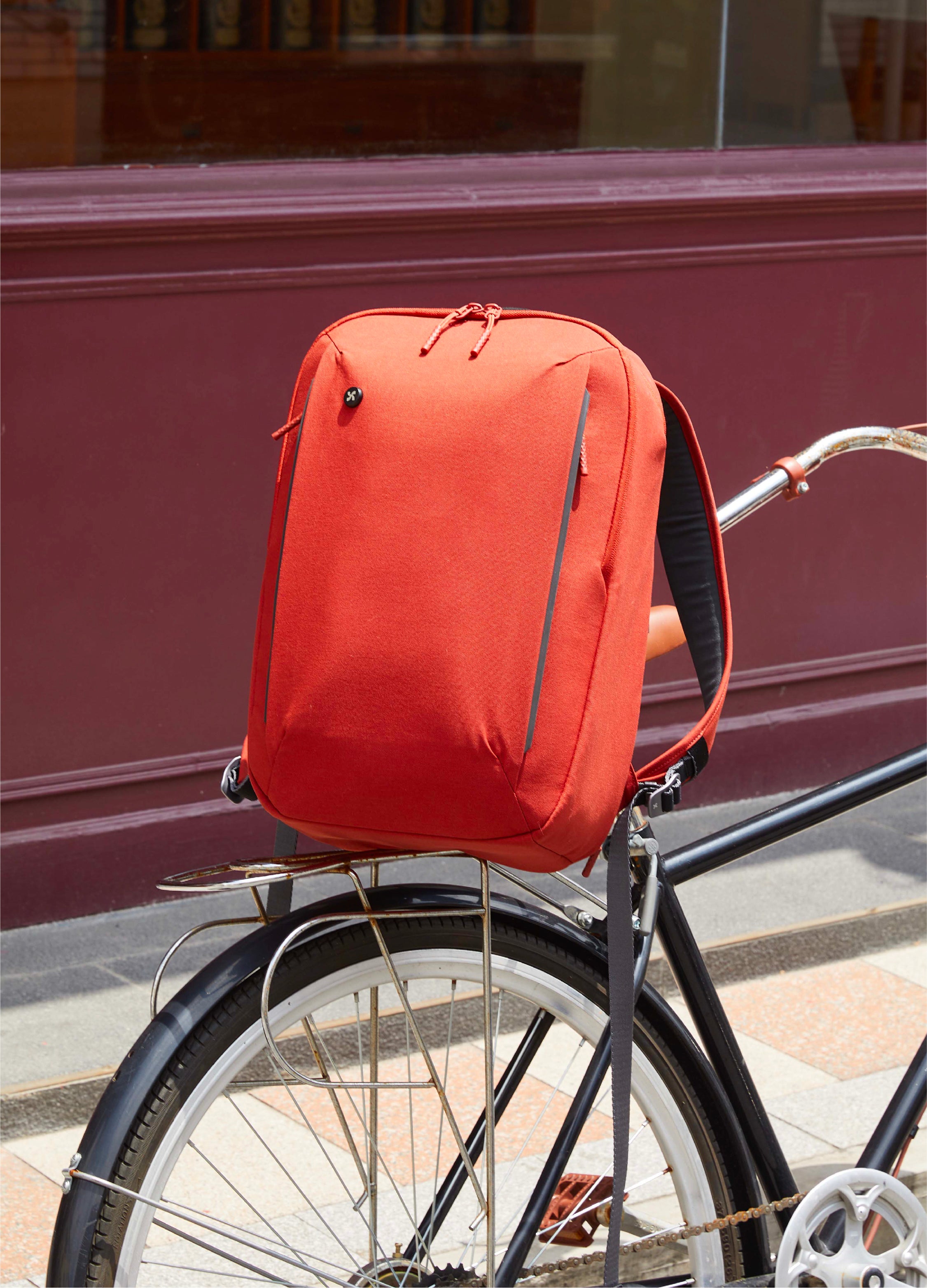 The Mopak City Adventure Backpack in bright red, casually placed on the rear rack of a city bicycle, highlighting its urban commuter-friendly design.