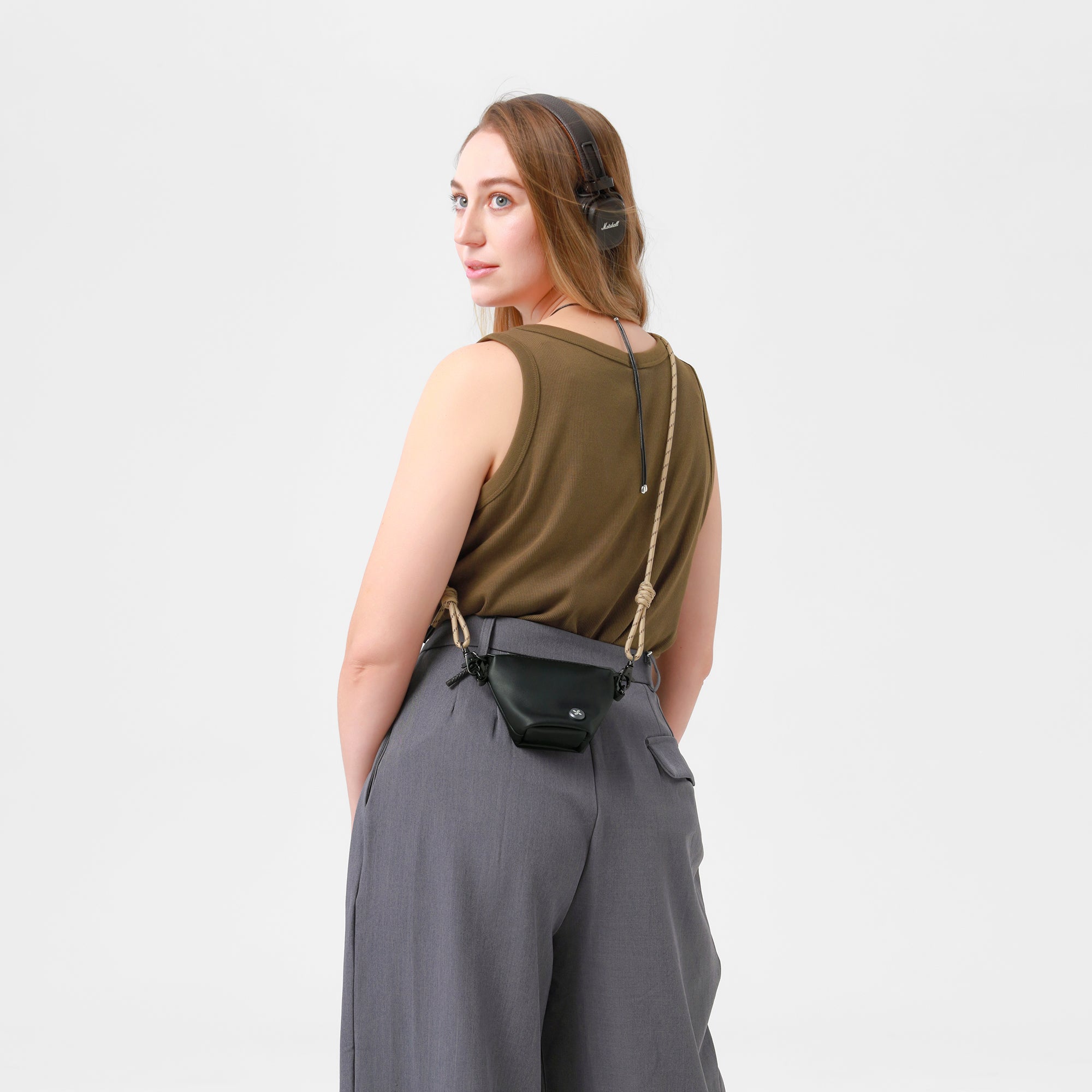 Woman wearing a green sleeveless top and gray pants with a black Kaf Mini bag on a light gray background