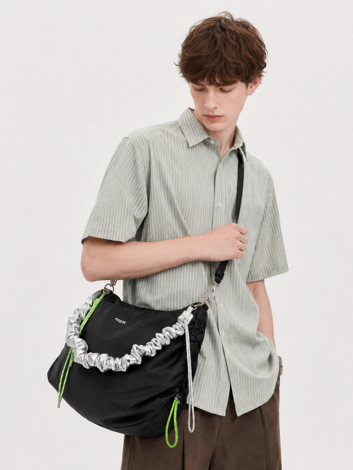 A model wearing a light green striped short-sleeve button-up shirt and brown trousers, carrying a black shoulder bag with a distinctive silver ruffled trim and bright green drawstring accents.