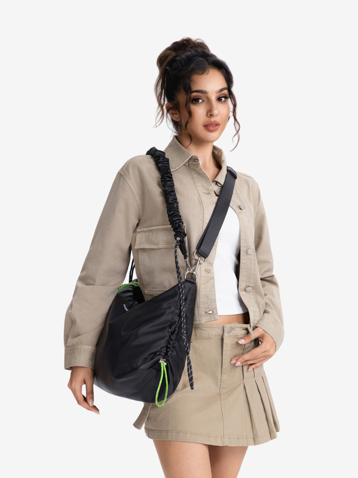 A woman models a black nylon shoulder bag with a black braided strap and a bright green zipper pull, styled with a beige utility jacket and matching pleated skirt against a light gray background.